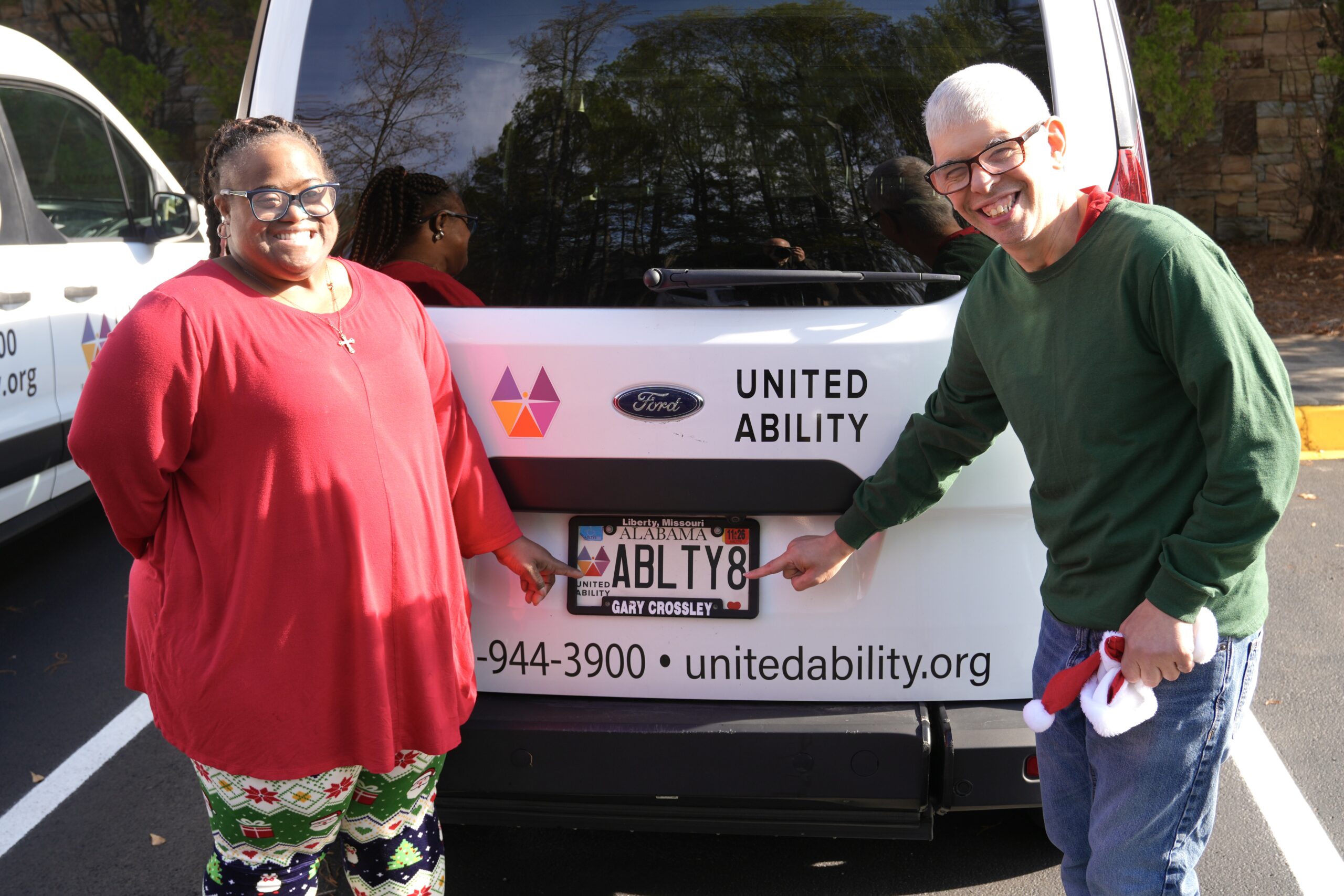 Two adults standing behing a United Ability van pointing at the United Ability specialty car tag.